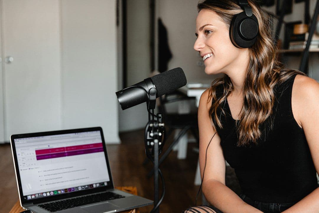 Podcast host in headphones smiling as she talks into a studio microphone, with an audio editing app open on a laptop beside her, the kind of episode an AI clip maker for podcasters turns into short clips